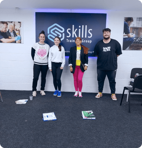 Four participants standing in a classroom during a First Aid Course session, with the 'Skills Training Group' logo on the wall behind them. The group is casually dressed, and course materials, including booklets and notebooks, are laid out on the carpeted floor. The walls feature posters of training scenarios.